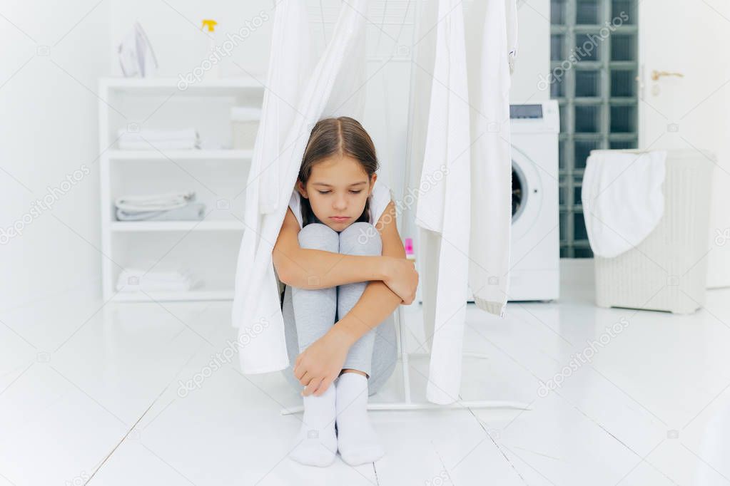 Small adorable girl sits on floor, being punished by parents, poses near clothes dryer, focused down with sad expression, washing machine, basket with laundry and console, thinks over something