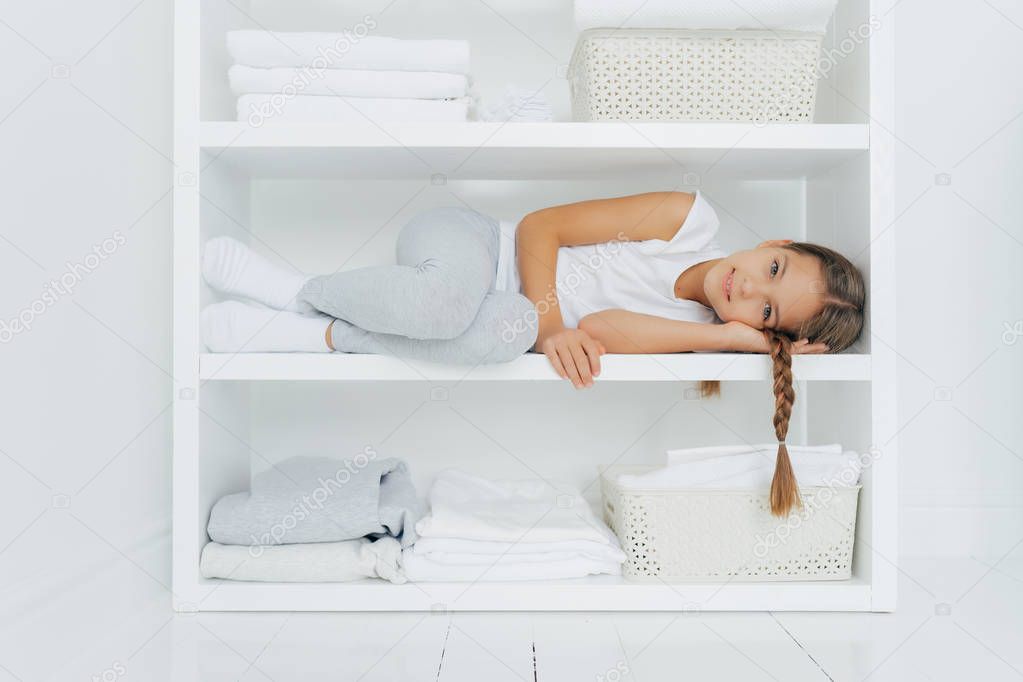 Horizontal shot of restful girl with pigtail lies on white console shelf, dressed in casual clothes, surrounded with piles of folded linen, basin of fresh laundry, has rest after helping mother