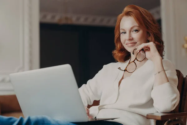 若い美しい赤毛の熟練した女性は 現代のラップトップを介して顧客にファイルを送信し 快適なアームチェアに座って アイウェアを保持し オンライン教育に時間を費やして ウェブセミナーを見て 喜んで式を持っています — ストック写真