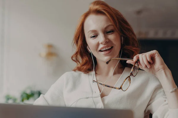 自信を持って満足した赤毛の女性は ラップトップコンピュータに集中し ラップトップコンピュータ上のビデオや映画を見て フレンドリーなビデオ通話を楽しんで 透明なガラスを保持し 無料のインターネット接続を使用しています — ストック写真