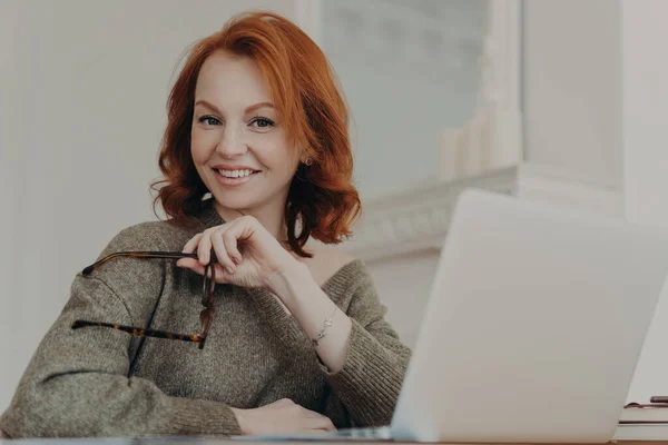 熟練した成功した赤毛の女性は ビジョン保護のためのアイウェアを保持し 開いたラップトップコンピュータの前に座って コワーキングスペースでポーズをとり セーターを着て 現代的なデバイスを使用しています 距離作業概念 — ストック写真