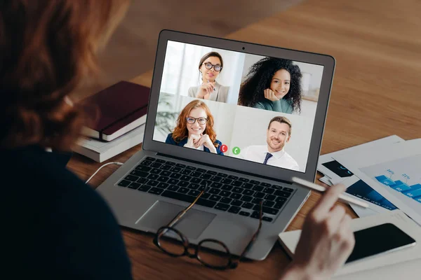 Businesswoman uses laptop application for communication with several people, has vitrual conference, works freelance from home, sits at desktop, surrounded with paper documents. Online negotiations