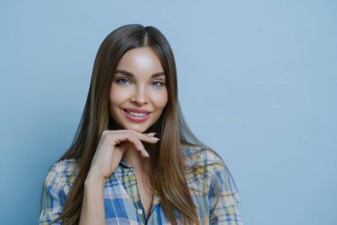 Sevimli, arkadaş canlısı bir bayanın yakın portresi çeneye dokunur, sağlıklı bir teni, asgari makyajı, sıcak bir ifadeyle, mavi arka planda güzel poz veren saçları vardır. Güzellik, cilt bakımı