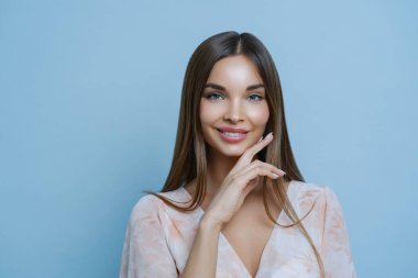 Güzel esmer kadının stüdyo portresi çeneye dokunur, güzelliğini gösterir, tenine değer verir, duyarlılığı hisseder, şık bir bluz giyer, mavi arka planda soyutlanır, rahat ve romantik bir ruh hali içindedir.