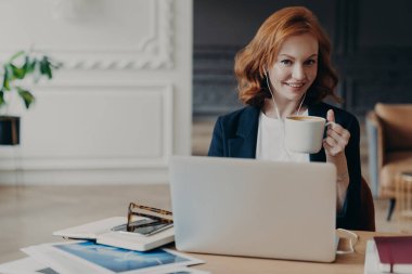 Mutlu, genç, kızıl saçlı bir kadın dizüstü bilgisayarda web sitesi eğitimi alıyor, proje bilgilerini tarıyor, kahve ya da espresso içiyor, masaüstünde poz veriyor, e-öğrenim için kulaklık kullanıyor, otomatik olarak aktifleştiriyor.