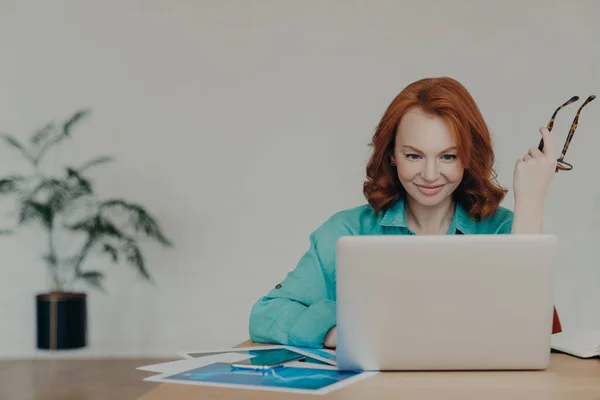 Happy beautiful female financial analyst prepares economic research, works on laptop computer, consults clients online, poses at desktop with documents, holds spectacles, busy with finishing task