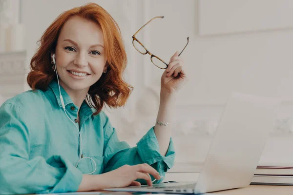 青いシャツの赤い髪の女性は ノートパソコン上で動作し ウェビナーやオンライン通信を見るためにイヤホンを使用し コースをリッスンし パンデミックコロナウイルス発生中に自宅からリモートで研究 — ストック写真