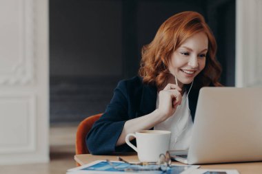 Mutlu gülümseyen kadın girişimci modern dizüstü bilgisayarla çevrimiçi konferans veriyor, evdeki kablosuz internete bağlanıyor, kulaklıkları daha iyi duymak için kullanıyor, birlikte çalışan alanda oturuyor.