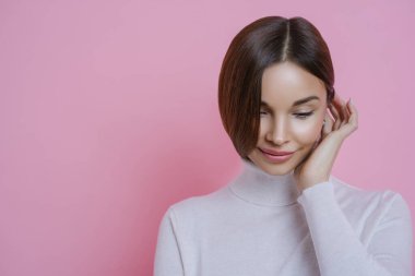 Düşünceli, utangaç genç bir kadının fotoğrafı aşağı bakıyor, rüya gibi bir ifadesi var, günlük kıyafetler giyiyor, hoş bir şeyler düşünüyor, pembe arka planda izole edilmiş, terfiniz için boş bir yer.