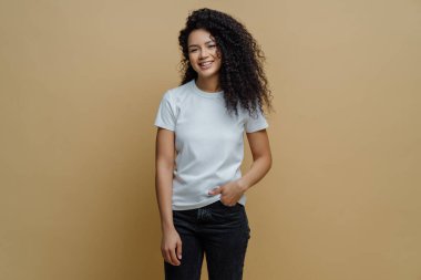 İnce, neşeli, kıvırcık saçlı, mutlu bir şekilde gülümseyen beyaz tişört ve kot pantolon giyen, cebinde ince vücutlu, bej arka planda izole edilmiş bir Afrikalı Amerikalı kadının fotoğrafı.