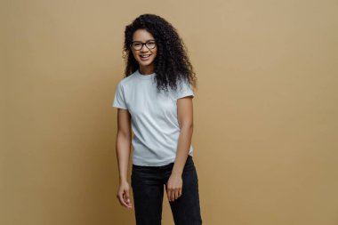 Gülümseyen esmer yüzlü genç bir kadın, optik gözlük takıyor, beyaz tişört ve kot pantolon giyiyor, bej arka planda tasasız poz veriyor. İnsanlar, mutlu ruh hali ve yaşam tarzı konsepti.