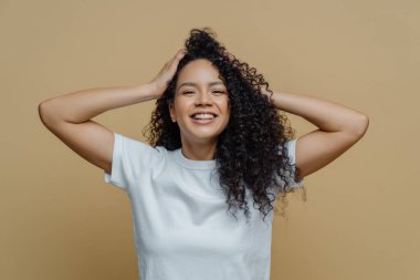 Happy curly woman keeps hands on head, has eyes full of happiness, smiles pleasantly and enjoys life, dressed in snow white t shirt, poses against beige studio background, glad to hear awesome news clipart