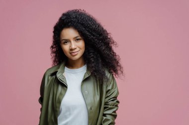 Ciddi koyu tenli bir kadının görüntüsü kameraya sakin bir ifadeyle bakar, çok dikkatli bir şekilde bir şeyler dinler, şık kıyafetler giyer, pembe stüdyo geçmişine karşı poz verir.