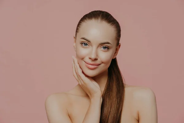 Portrait of good looking female has long straight hair combed in pony tail, stands shirtless, has clean perfect skin, satisfied with her natural beauty, poses indoor against pink background.