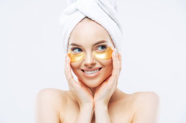 Taze ve sağlıklı cilde sahip genç bir kadın, gözlerinin altına altın bantlar takıyor, koyu halkalar üzerinde mükemmel kozmetik ürünler sergiliyor, gömleksiz duruyor, beyaz arka planda izole edilmiş..