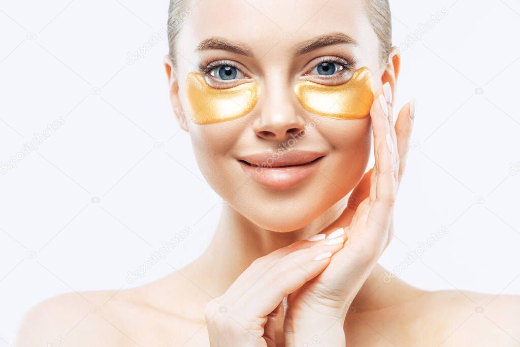 Close up shot of charming tender young woman applies gold collagen patches on fresh facial skin, touches face, has makeup, well cared body, stands against white background. Under eye treatment.