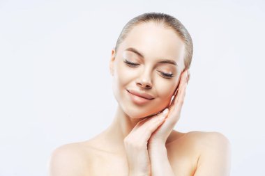 Şefkatli güzel bir kadının portresi yanağa dokunur ve içeride çıplak durur, günlük hijyen prosedüründen zevk alır, aşağıya bakar, tenini önemser, romantik bir ruh halinde olur, beyaz stüdyo duvarının üzerinde durur..