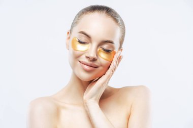 Koyu renk saçlı güzel bir kadın, gözlerini kapatır ve yüzüne dokunur, altın kolajen bantlar takar, kırışıklık önleyici maskeyi kaldırır, çıplak omuzlarını kaldırır, beyaz arka planda durur. Spa tedavisi