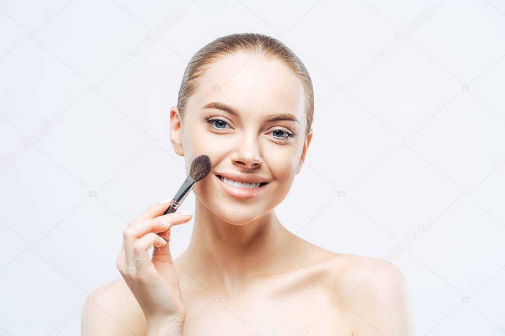Adorable young brunette woman applies cosmetic tonal foundation with beauty brush, smiles tenderly, has healthy skin, well cared body, isolated on white background. Women, skin care, makeup concept