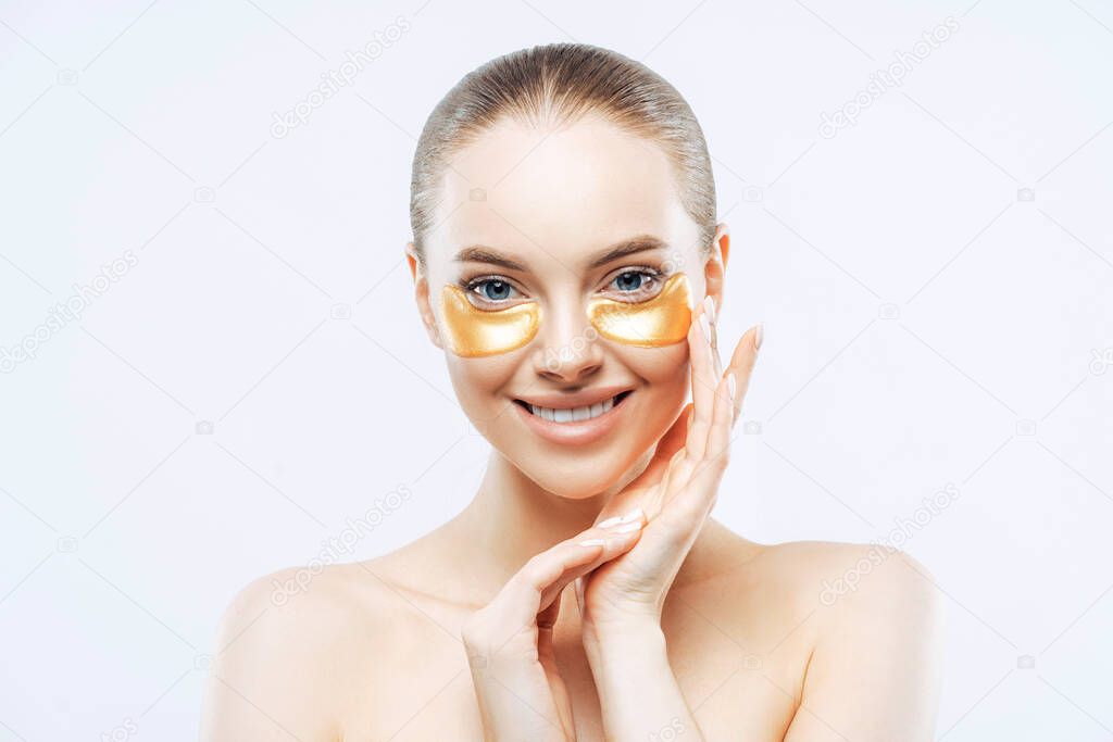 Studio shot of attractive European woman touches face gently, smiles tenderly, applies golden hydrogel patches under eyes, isolated on white background. Eye mask treatment. Anti age procedure