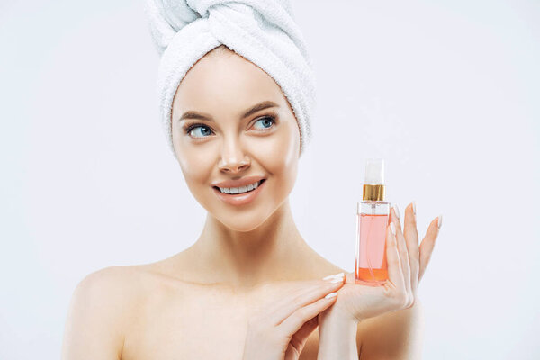 Beauty portrait of happy smiling European woman with healthy skin, makeup, holds bottle of aromatic parfume, stands naked indoor, wears towel on head. Women, cosmetology and skin care concept