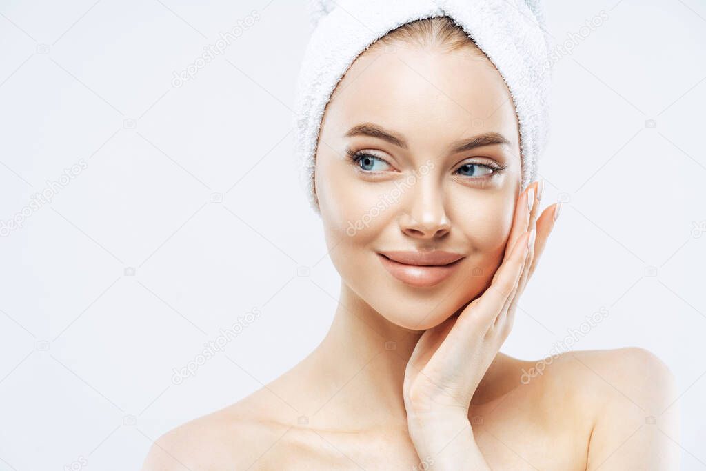 Beautiful female model with makeup, touches cheek, looks aside, has fresh clean skin after taking bath, wears soft towel on head, isolated over white background, copy space. Face care, hygiene concept