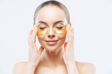Güzel gülümseyen Avrupalı bir kadının yakın portresi gözlerini kapatıyor, yaşlanmayı önleyen göz terapisinden hoşlanıyor, altın bantlar takıyor, temiz bir cildi var, beyaz arka planda üstsüz duruyor. Yüz bakımı
