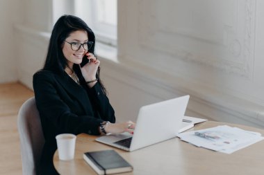 Güzel kadın yönetici uzaktan iş yapıyor, cep telefonuyla konuşuyor ve iş yerindeki bilgisayardaki bilgileri kontrol ediyor. Etrafı kahve kağıdı kağıtlarıyla çevrili. Not defterinde telefon görüşmeleri var.