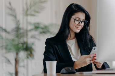 Tatmin olmuş iş kadını akıllı telefon kullanıyor, bilgisayar ekranının önünde iş yerinde poz veriyor, bir fincan lezzetli içecek molada mesajlar gönderiyor, çağdaş ofiste poz veriyor. Teknoloji ve iş kavramı