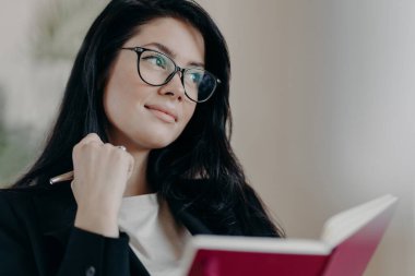 Düşünceli kadın işletme sahibi planlama programı üzerinde çalışıyor, not defterine yazıyor, kalemini tutuyor, düşünceli bir şekilde bakıyor, resmi kıyafetler ve gözlük takıyor, yaratıcı fikirler düşünüyor, bir kenara konsantre oluyor.