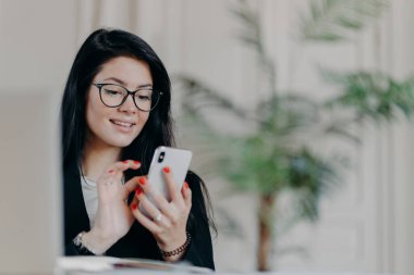 Mutlu esmer iş kadını akıllı telefon kullanıyor, gözlük takıyor, ortağına mesaj gönderiyor, proje üzerinde çalışıyor, iş yerindeki samimi iç mekanlara karşı poz veriyor. Teknoloji, işgal ve iş kavramı