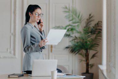 Ciddi esmer kadın çalışan masanın yanında duruyor, elinde kağıt belgeler var, akıllı telefonla konuşuyor, resmi kıyafetler ve gözlük takıyor, iş stratejisini partneriyle tartışıyor, toplantı düzenliyor.