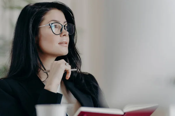 Thoughtful European woman makes notes in notepad, holds pen and writes down some ideas, makes list of goals, plans day in personal organizer, wears transparent glasses, thinks about something