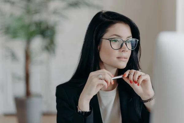 Skilled female freelancer concentrated at computer display, wears optical glasses, holds pen, has dark hair, makes online banking. Business lady works distantly from home, develops new startup project