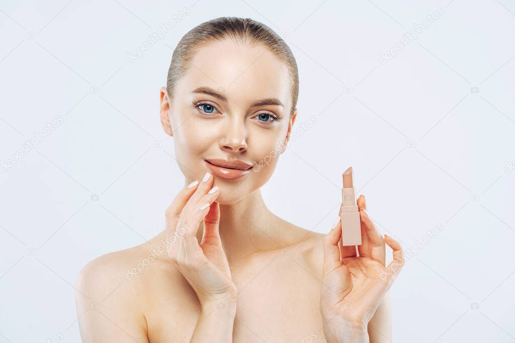 Isolated shot of beautiful girl applies lipstick on lips, has perfect makeup, poses with beauty product, touches face gently, poses shirtless, gets ready for work, poses against white background