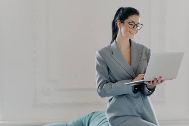 Dizüstü bilgisayardaki mutlu kadın girişimci tipler, iş merkezindeki modern çalışma ortamında çalışıyor, resmi kıyafetler giyiyor, internette sörf yapıyor, Webinar eğitimi izliyor, partnerlerle online sohbetler yapıyor.