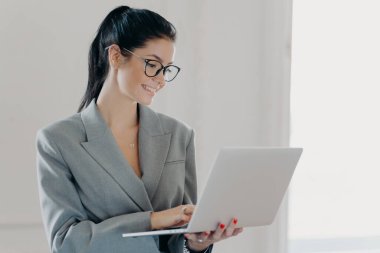 Kendine güveni tam bir ekonomist kadın şirket gelirini laptopta analiz ediyor, multimedya dosyalarını paylaşıyor, at kuyruğu ile taranmış koyu renk saçları var, resmi kıyafetler giymiş, kapalı mekanda duruyor.