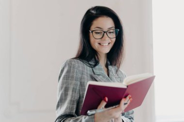 Yaratıcı, neşeli yazarın fotoğrafı ilginç bir makale yazmayı düşünüyor, yeni planlar yapıyor, mutlu bir şekilde gülümsüyor, gözlük takıyor, not defterine notlar yazıyor, resmi kıyafetler giyiyor.