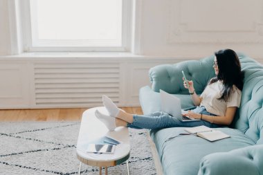 Rahat kız öğrencilerin yan fotoğrafları ev ödevi yaparken modern teknolojileri kullanır, proje yapmak için web kaynaklarını kullanır, rahat bir koltuğa oturur, masaüstüne yaslanır.