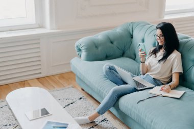Samrtphone ekranına odaklanmış güzel esmer bir kadının resmi. Gerekli bilgileri not defterine yazıyor. Webinar eğitimi izliyor, rahat koltukta rahat bir odada poz veriyor.