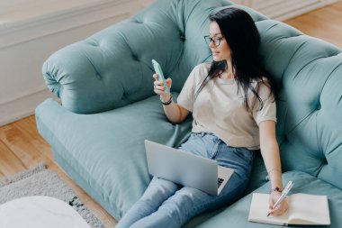 Akıllı telefon ekranına odaklanmış serbest çalışan meşgul kadın fotoğrafçı, internetteki bilgileri kontrol ediyor, günlüğe bir şeyler yazıyor, modern teknolojileri iş için kullanıyor, rahat bir odada poz veriyor.