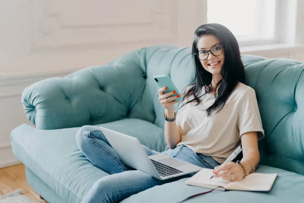 Glad female student works on course paper, surrounded with modern gadget, makes notes in notepad, feels relaxed at comfortable sofa, works online, wears casual clothes, watches video online.