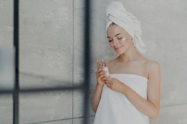 Tatmin olmuş genç beyaz kadın güzelliğine önem veriyor, yüzüne losyon kremi sürüyor, banyo havlusuna sarılı duruyor, ev tuvaletinde gri duvara karşı poz veriyor. Günlük ev bakımı ve kaplıca konsepti