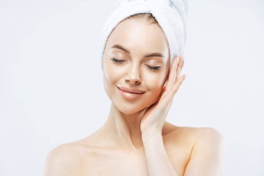 Kafasına havlu sarılmış, gözlerini kapatan, cildinin yumuşaklığından hoşlanan, cilt bakımından hoşlanan, çıplak omuzlar içinde duran, banyodan sonra dinlenen, beyaz arka planda izole edilmiş oldukça sakin bir kadın.