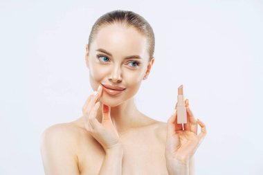 Dudak bakımı konsepti. Koyu renk saçlı, kozmetik ürünlerle poz veren, duygusallığı bir kenara bırakılmış, üstsüz poz veren, bakımlı, beyaz arka planda izole edilmiş güzel bir kadın.