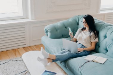 Serbest çalışan bir kadının yatay çekimi internet üzerinden para kazanıyor, akıllı telefondan e-postaları kontrol ediyor, modern teknolojilerle çevrili, not almak için organizatör, yüksek hızlı internet kullanıyor, rahat bir koltukta oturuyor.