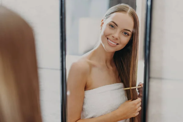 快乐美丽的年轻女子在镜子中看到自己的倒影 有着精心呵护的长发 用毛刷 用浴巾包裹 做发型 发型和自我护理概念 — 图库照片