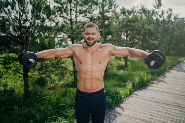 Formda yetişkin bir adam antrenman sırasında ağırlık kaldırır, çıplak gövdeyle poz verir, kaslı bir vücudu vardır, gücünü gösterir, halterleri kaldırır. Sokakta motivasyonlu vücut geliştirici