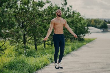 Kalın sakallı sporcu adam atlama halatıyla egzersiz yapıyor, kaslı gövdesi var, fiziksel olarak iyi durumda, dışarıda poz veriyor. Fitness ve sağlıklı konsept. Erkek koşucu spor malzemeleriyle ısınıyor