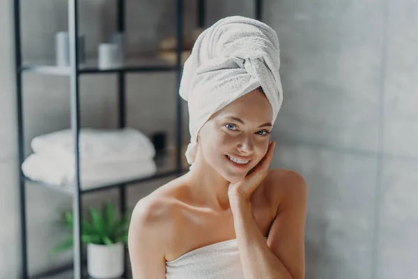 Beautiful European woman with makeup touches skin, has minimal makeup, has healthy glowing skin, wrapped in bath towel, enjoys rest at home. Spa woman poses in bathroom. Beauty, wellness concept